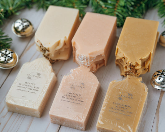 Three bars of handcrafted soap positioned side by side, each with a different color and label, against a background with Christmas decorations.